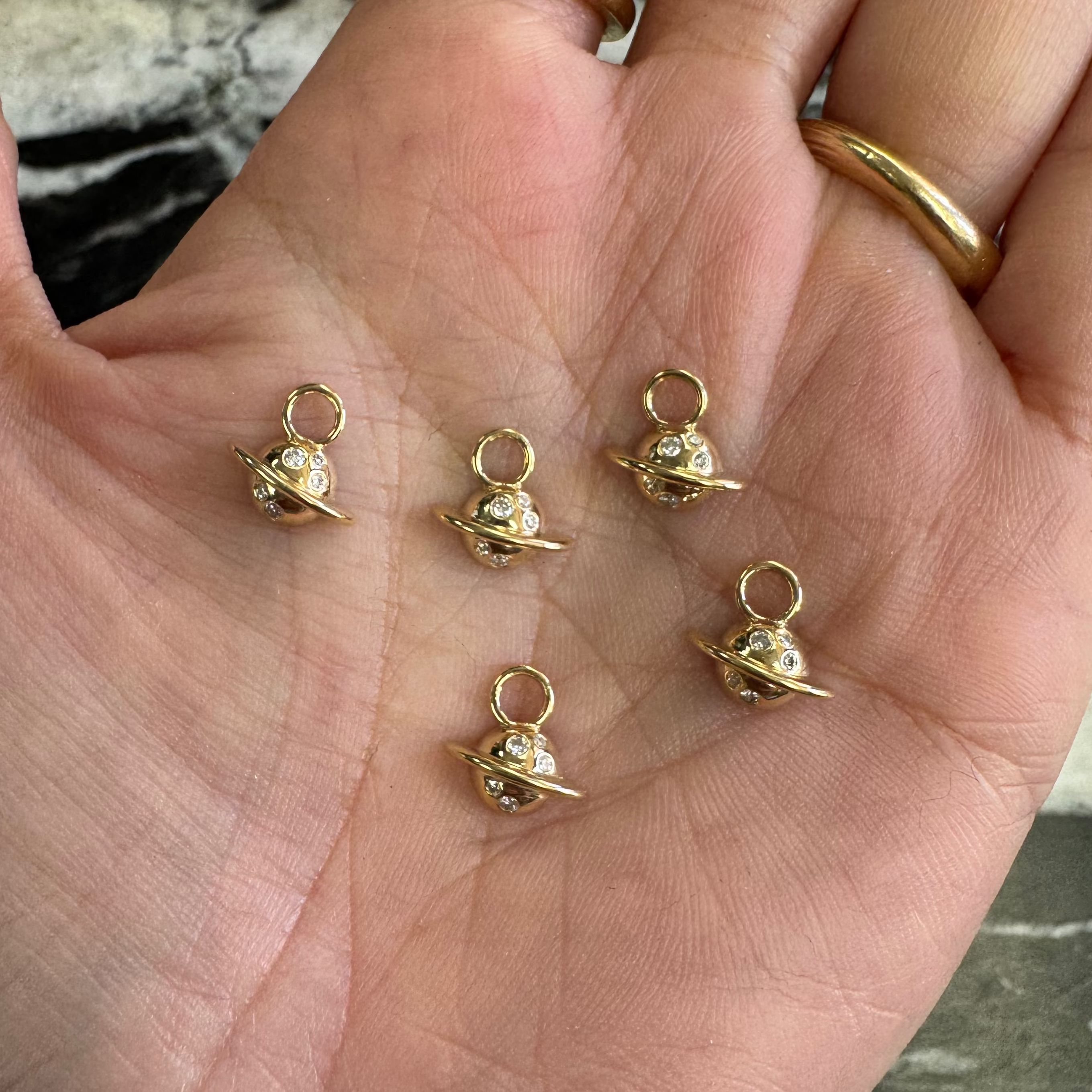 Five small gold planet charms with diamonds resting on an open palm, with two gold rings worn on the fingers.