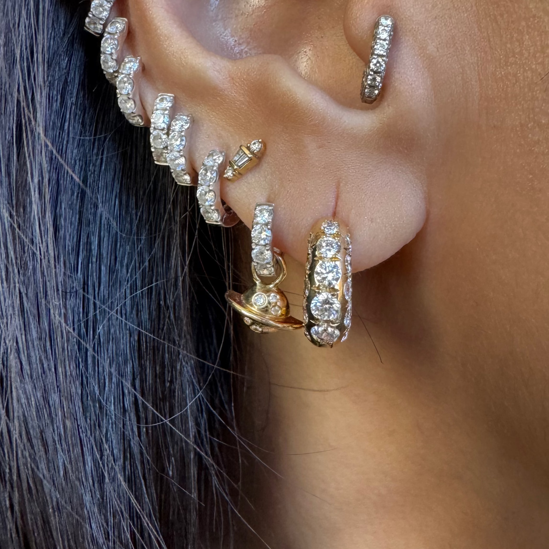 A close-up of a person's ear adorned with multiple gold and silver-toned huggie hoop earrings, some embellished with diamonds, shown against dark hair.