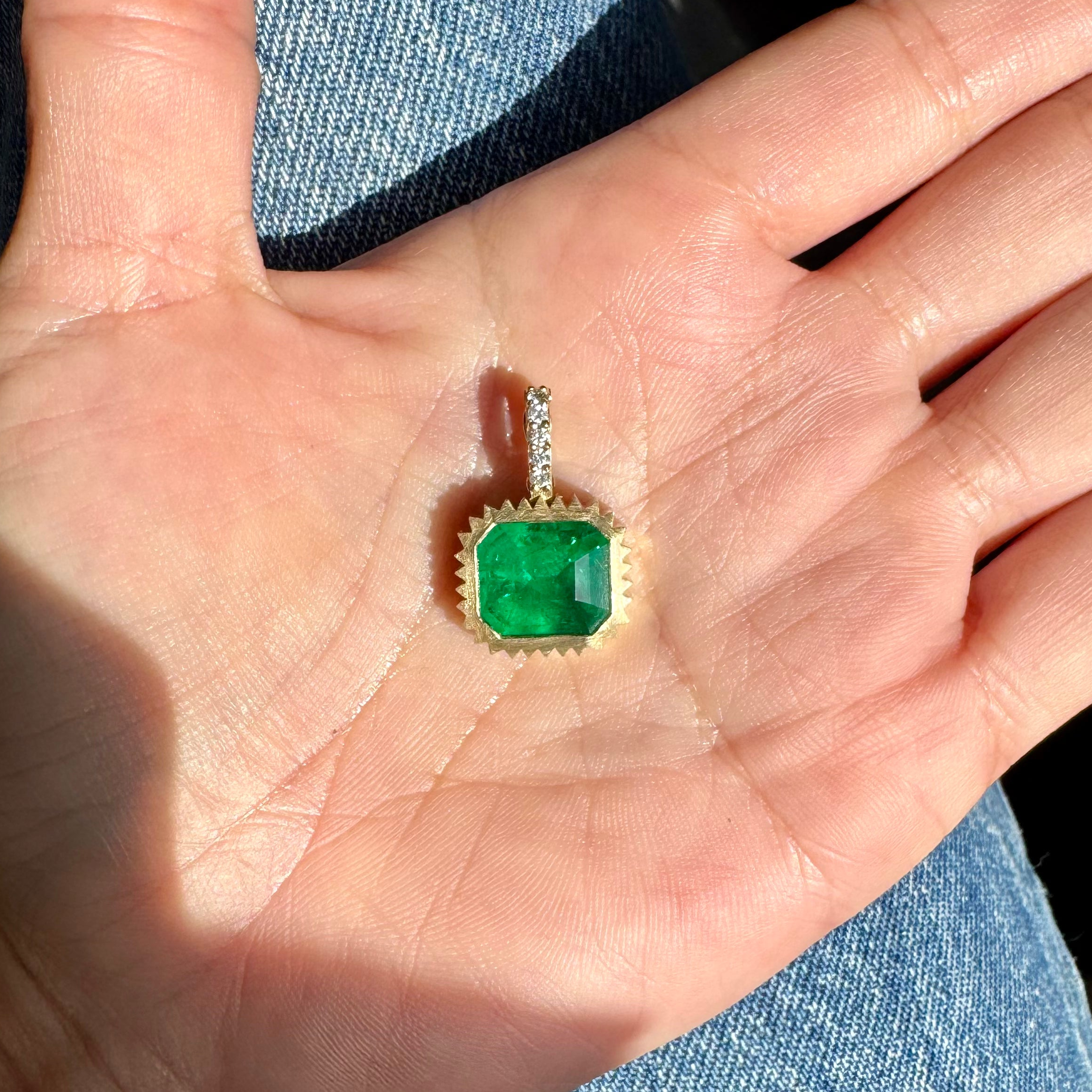 hand holding a gold pendant with an emerald-cut emerald and a diamond bail against a blue denim background 