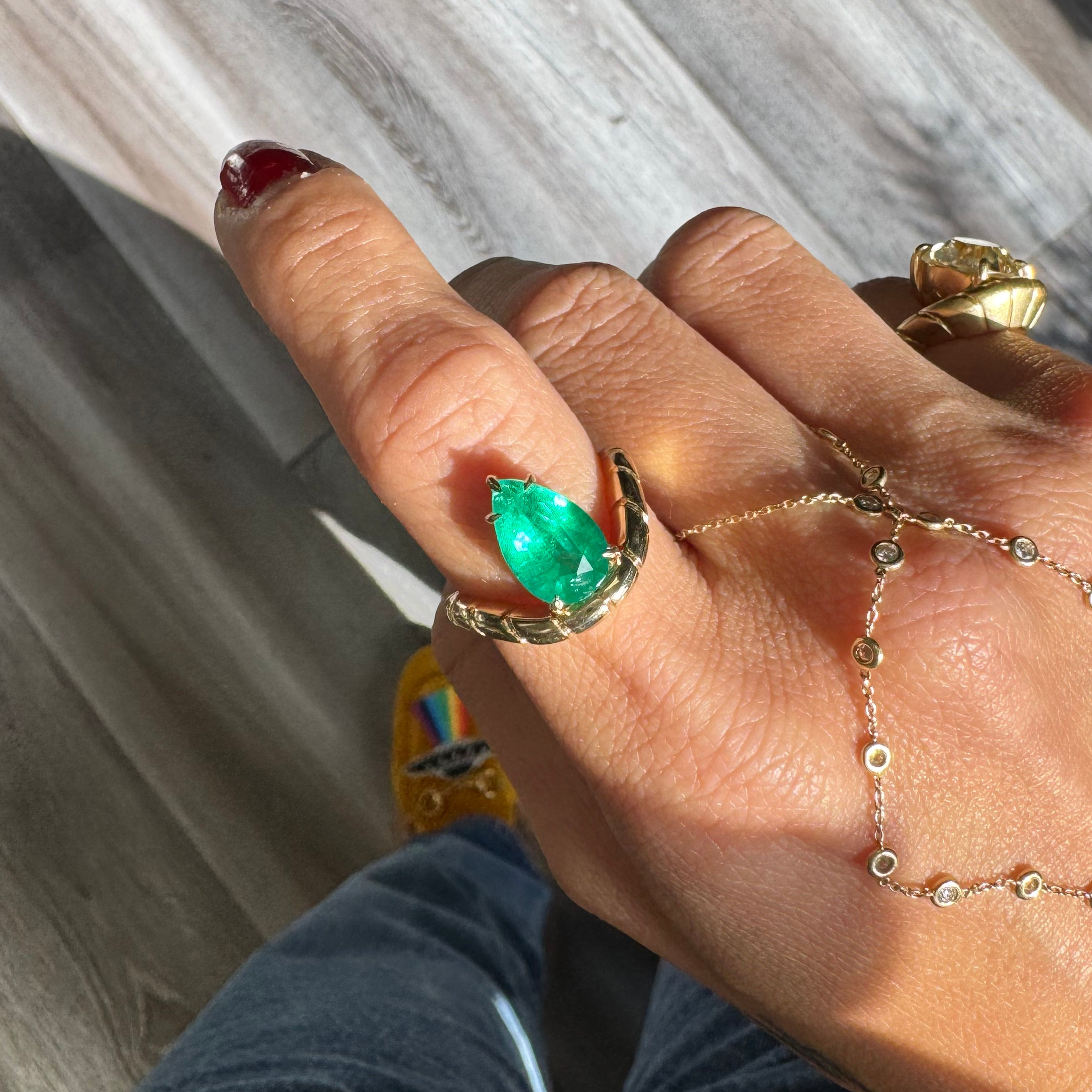 Person wearing an Emerald ring with a curved gold band against a multicolored background 