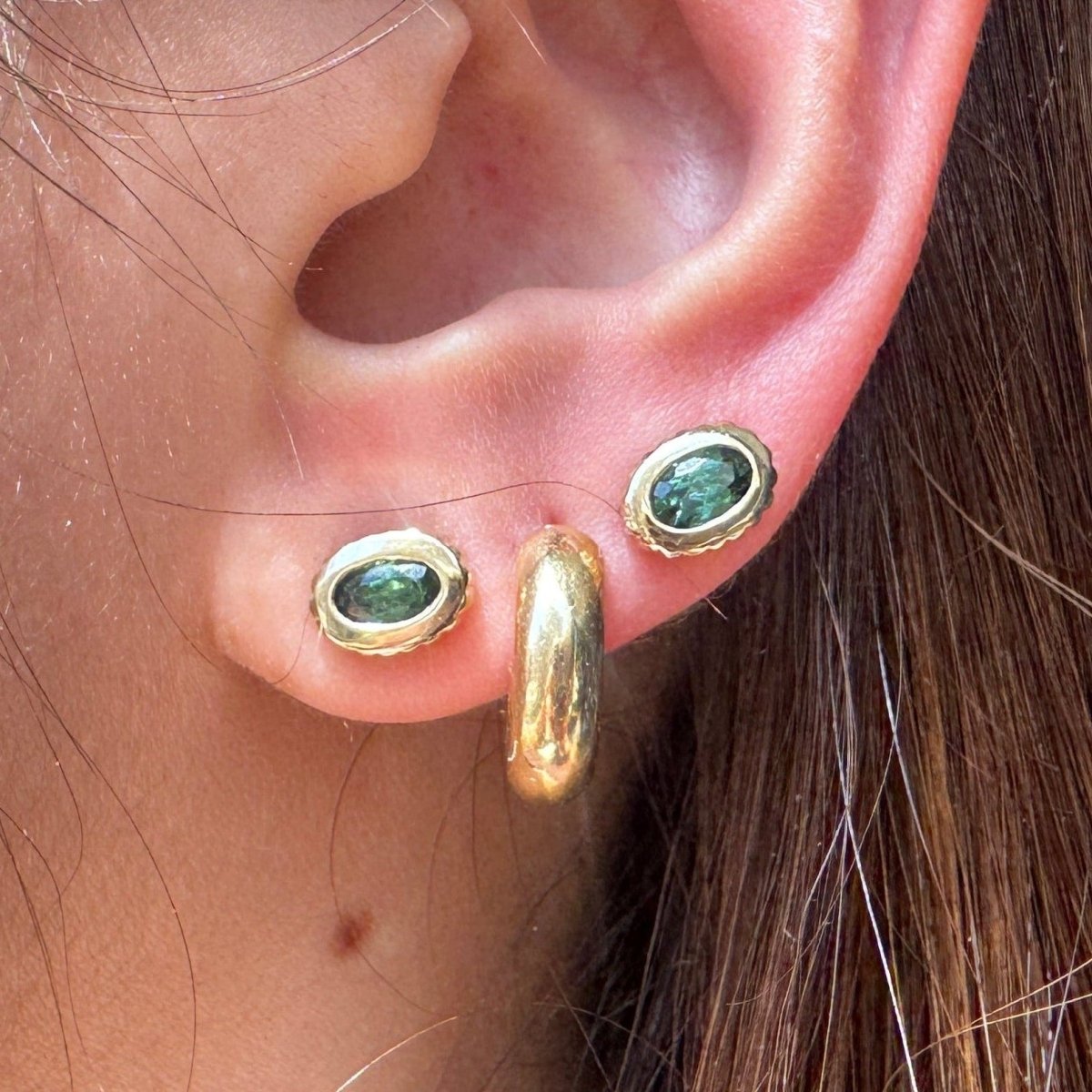 Ear wearing multiple gold earrings including gold studs with oval green tourmalines 