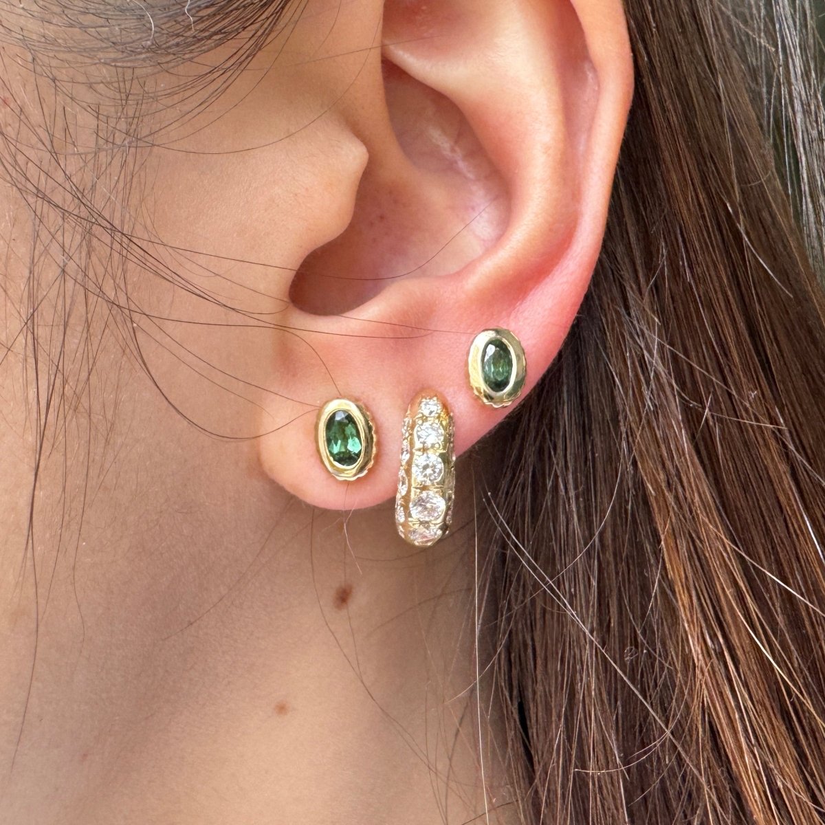Ear wearing multiple gold earrings including gold studs with oval green tourmalines 