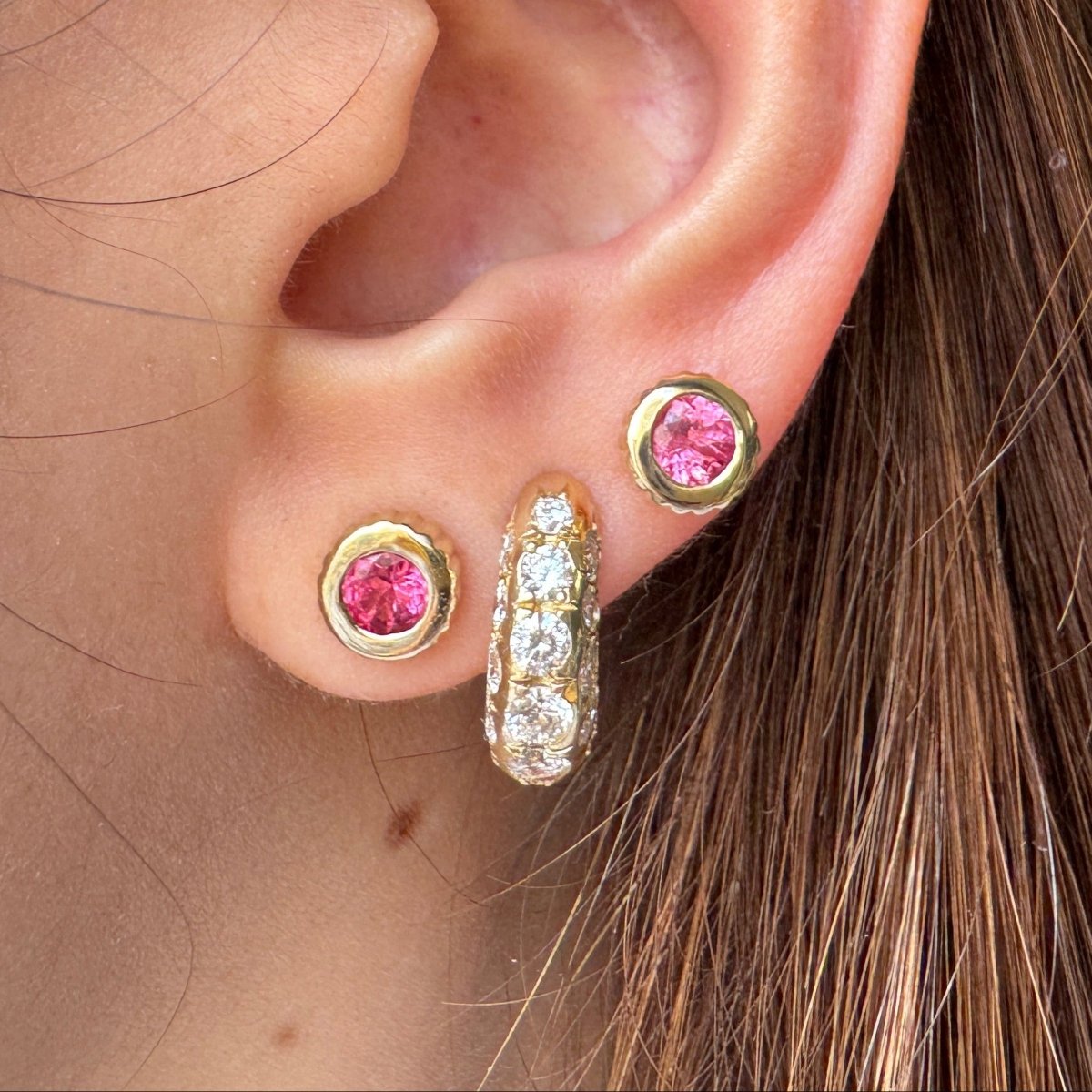 Ear wearing multiple gold earrings including two gold studs with round pink spinels 