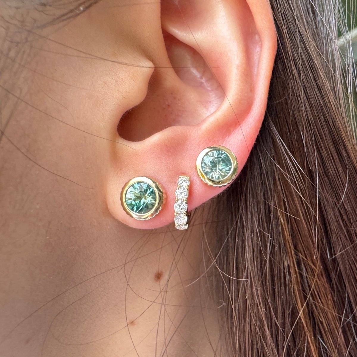 Ear wearing multiple gold earrings including two gold studs with round blue zircons