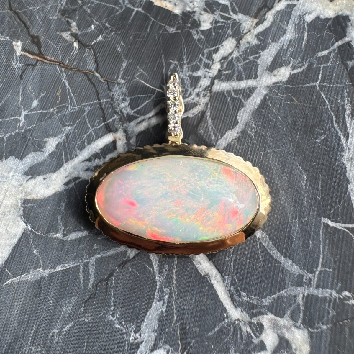 Gold pendant with a rainbow cabochon opal and a diamond bail against a black and white marble background