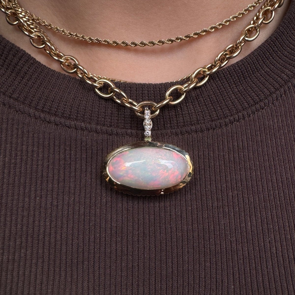 Person wearing a gold pendant with a rainbow cabochon opal and a diamond bail against a brown top