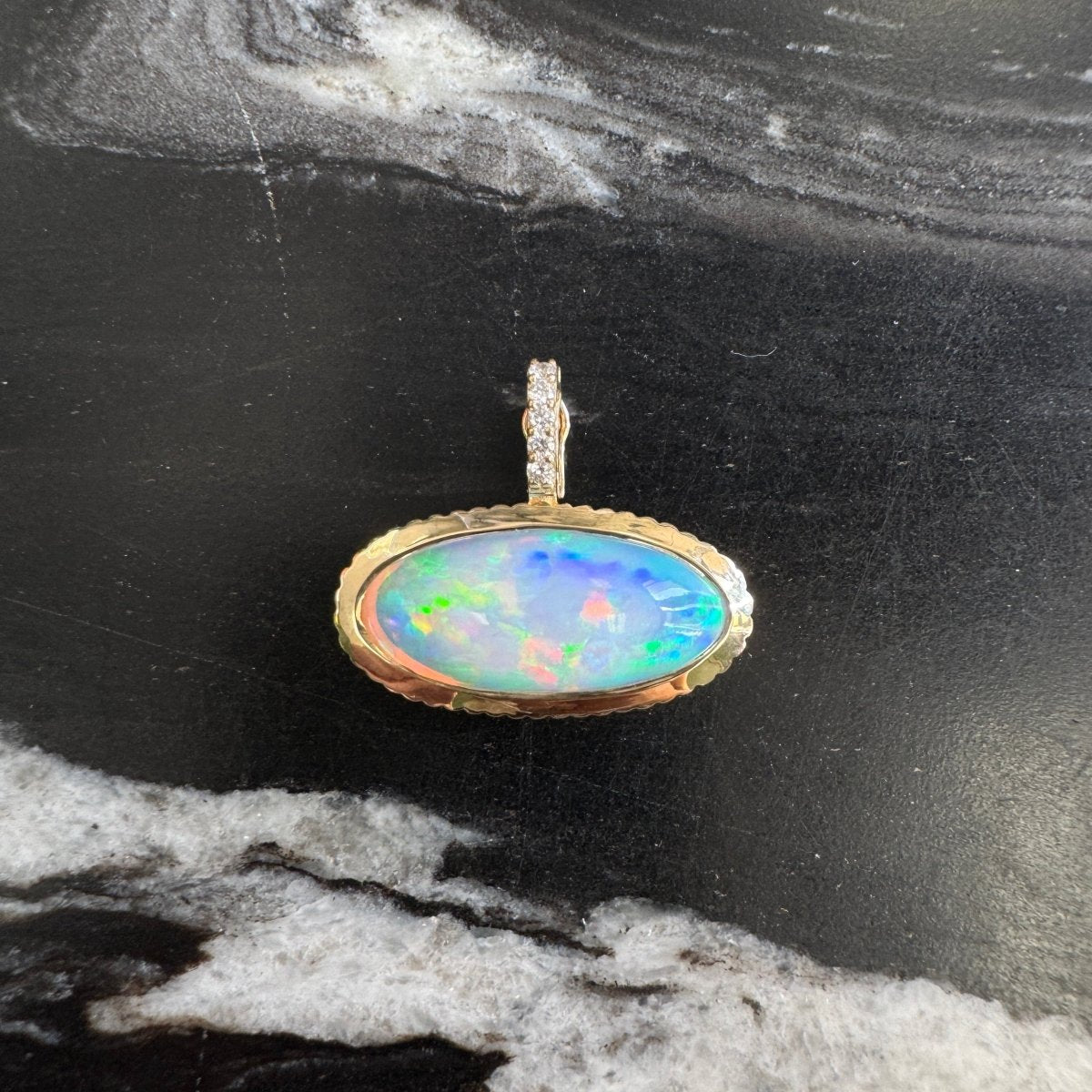 Gold pendant with a multicolored opal with a diamond bail against a black and white marble background