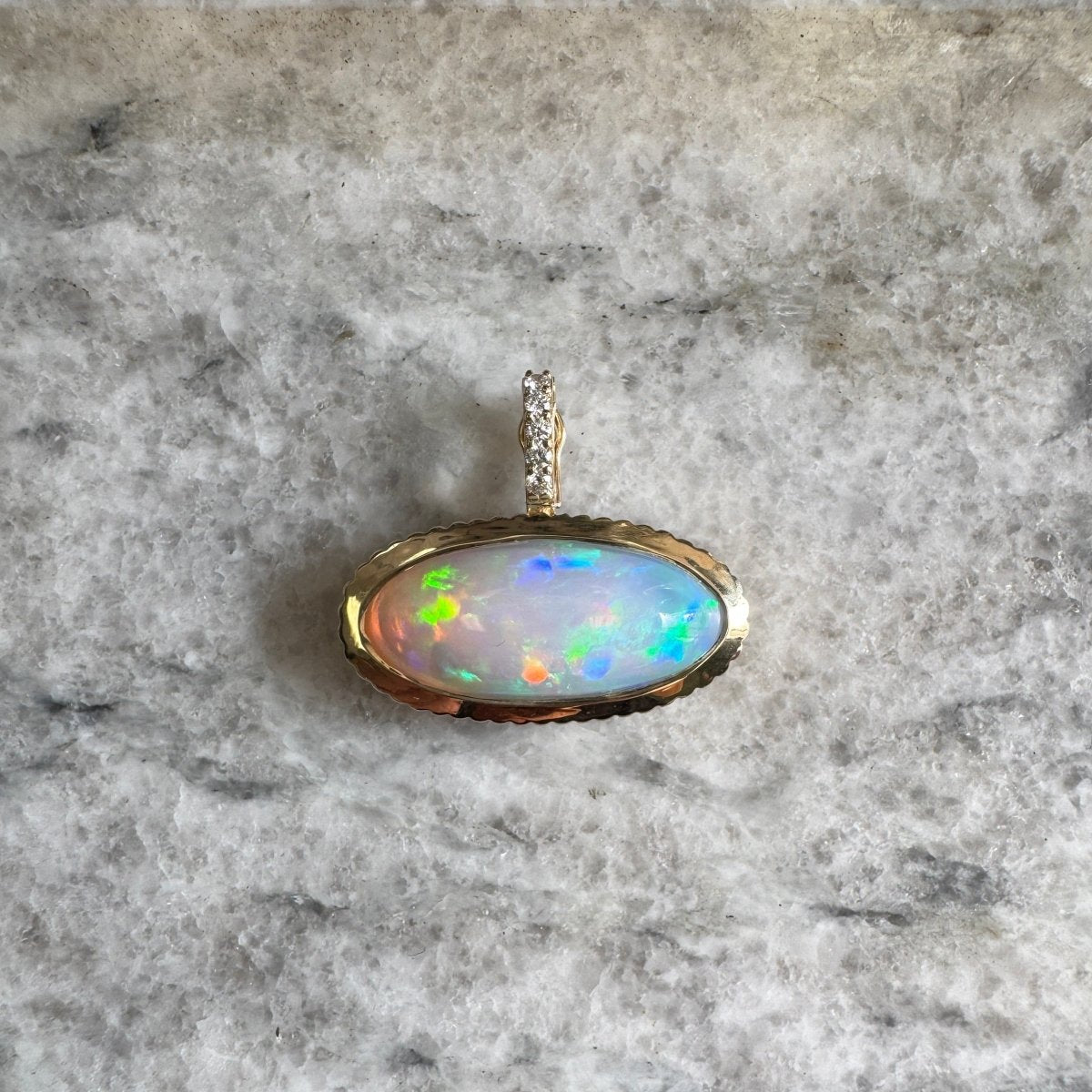 Gold pendant with a multicolored opal with a diamond bail against a black and white marble background
