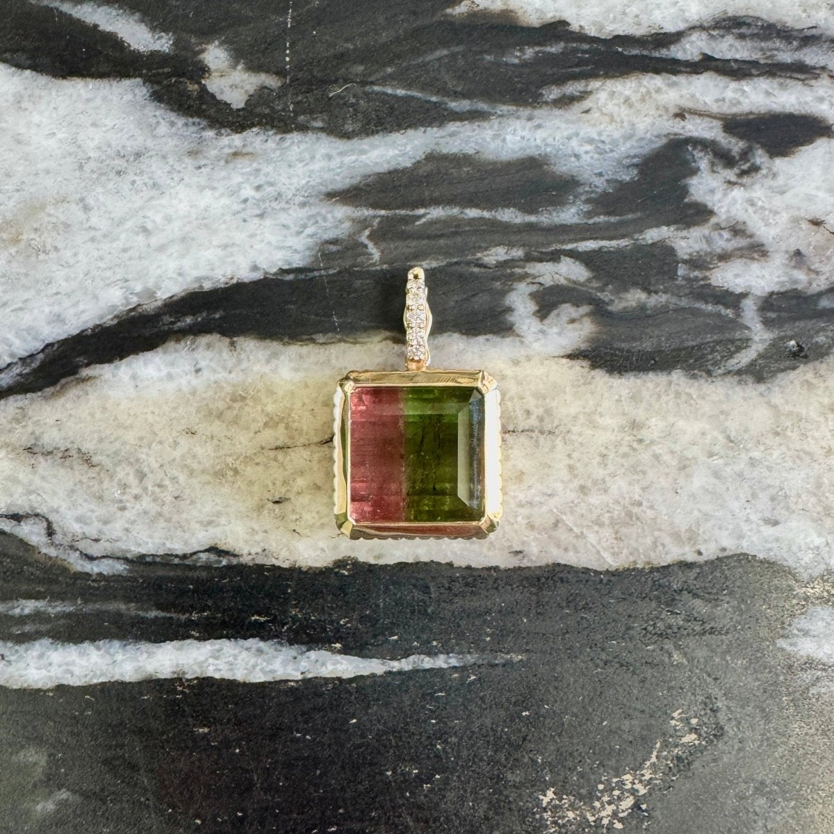 A square pendant with a gold-toned bezel setting featuring a green and pink banded stone, displayed on a black and white marbled surface.