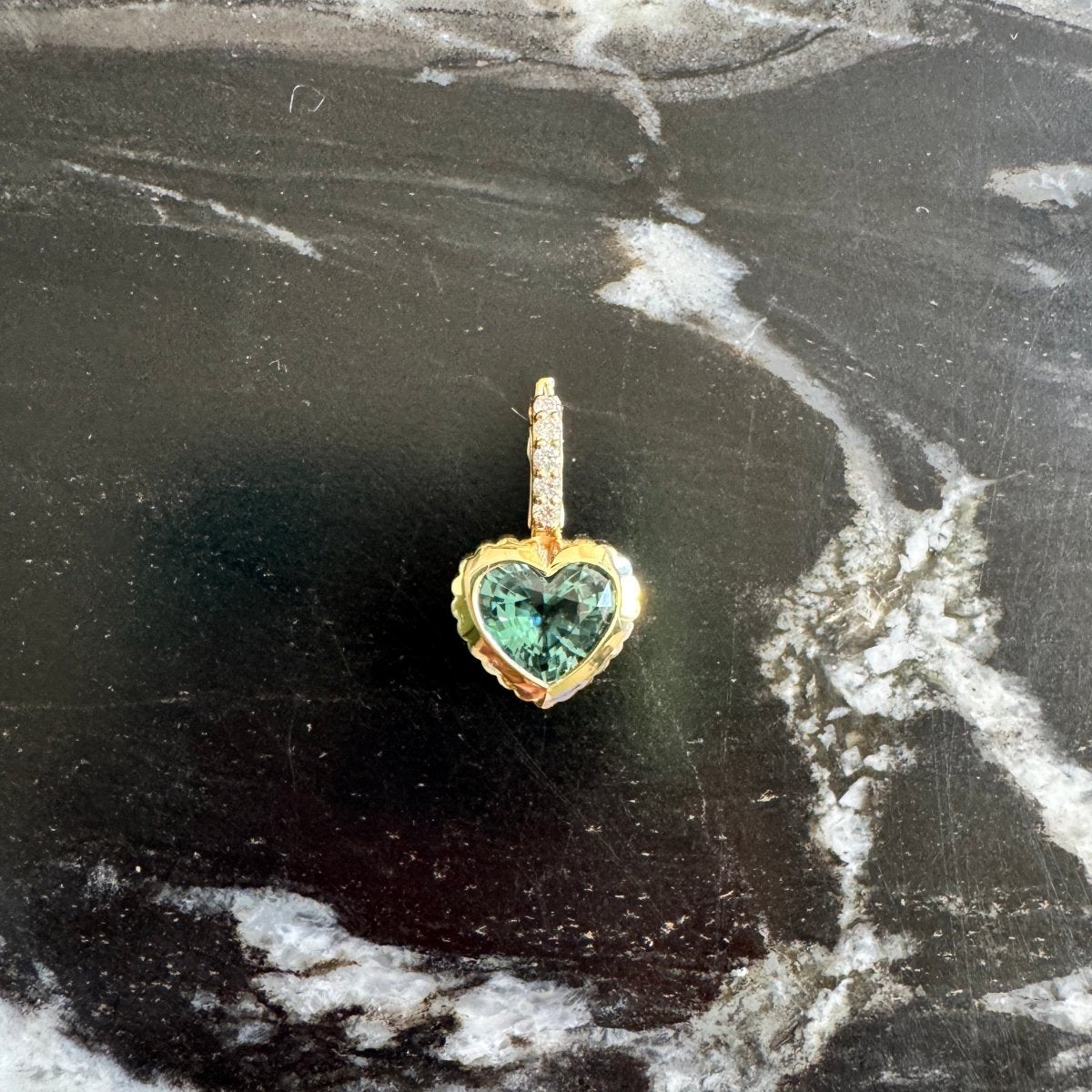 A small gold heart-shaped pendant with a green stone, resting on a black and white marbled surface.