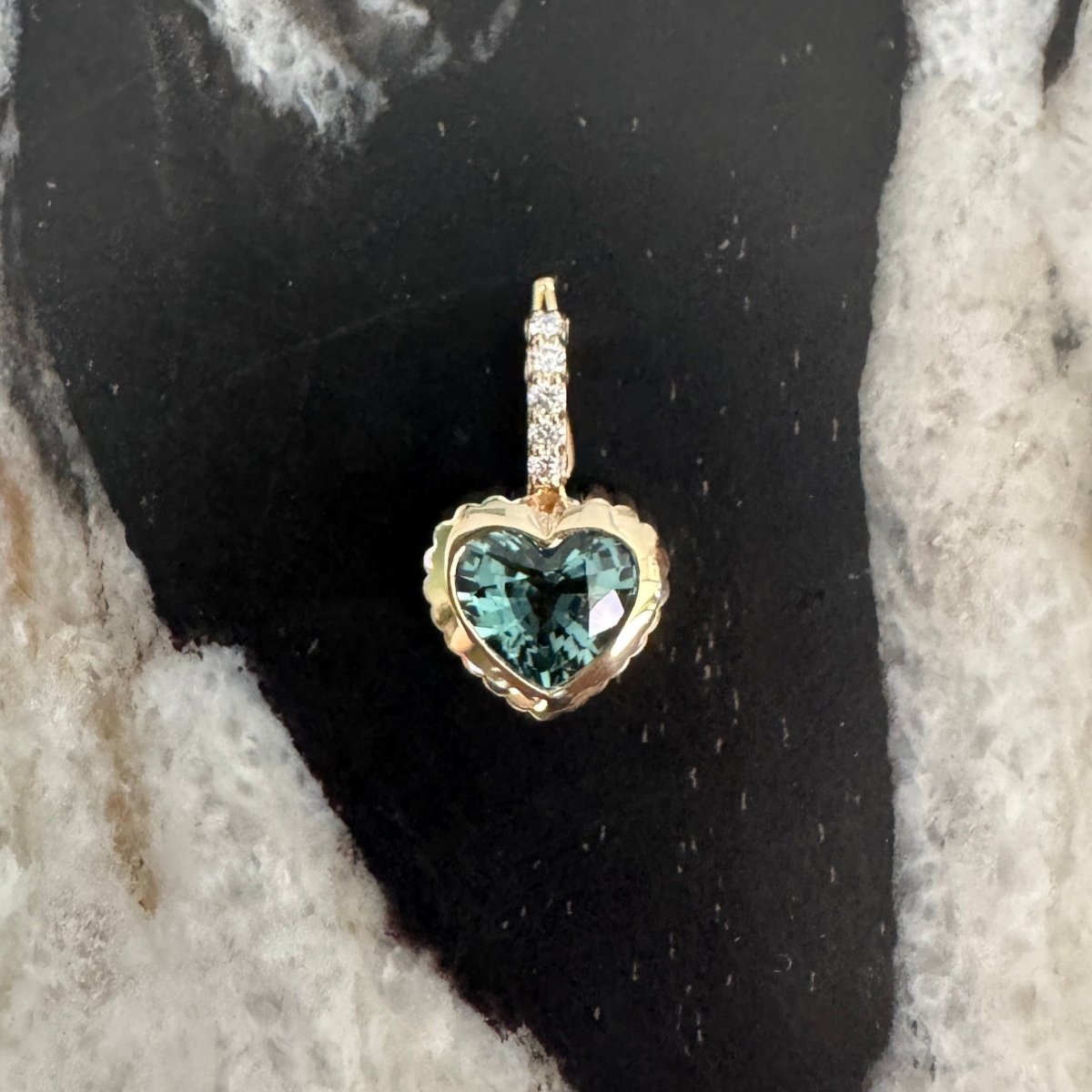 Gold pendant with a heart-shaped blue tourmaline and a diamond bail against a black and white marble background 