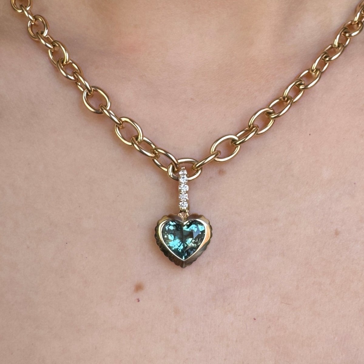 Person wearing a gold pendant with a heart-shaped blue tourmaline and a diamond bail on a gold chain