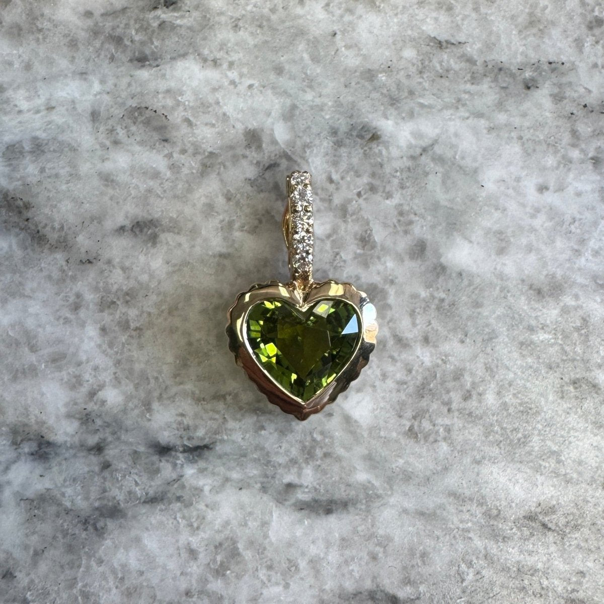 Gold pendant with a heart-shaped green tourmaline and a diamond bail against a black an white marble background 