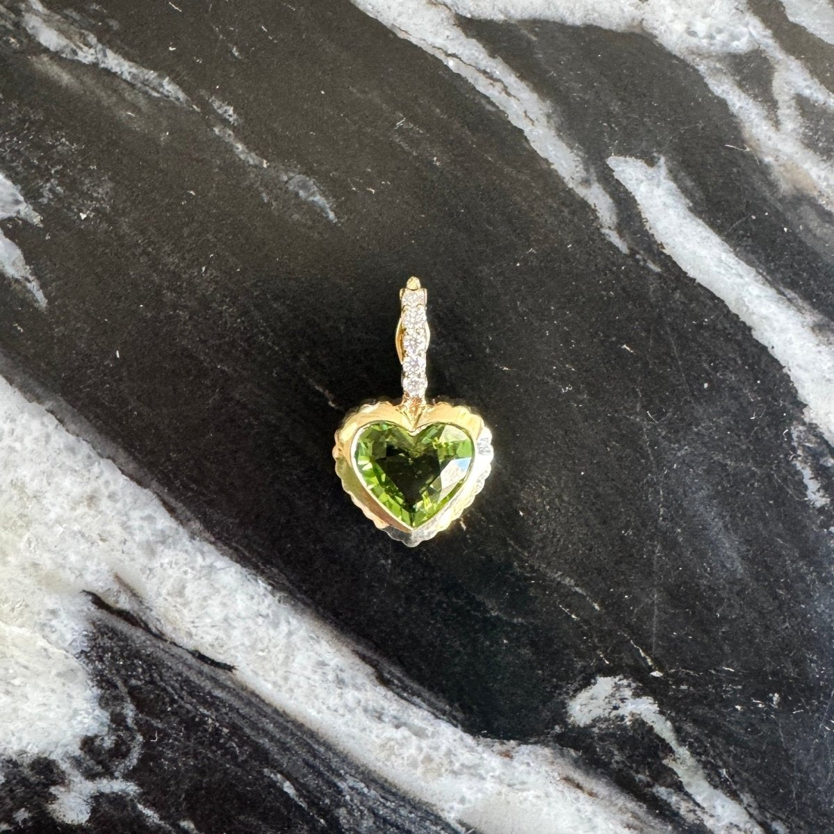 A small heart-shaped pendant with a green gemstone and gold-tone setting, photographed on a black and white marbled surface.