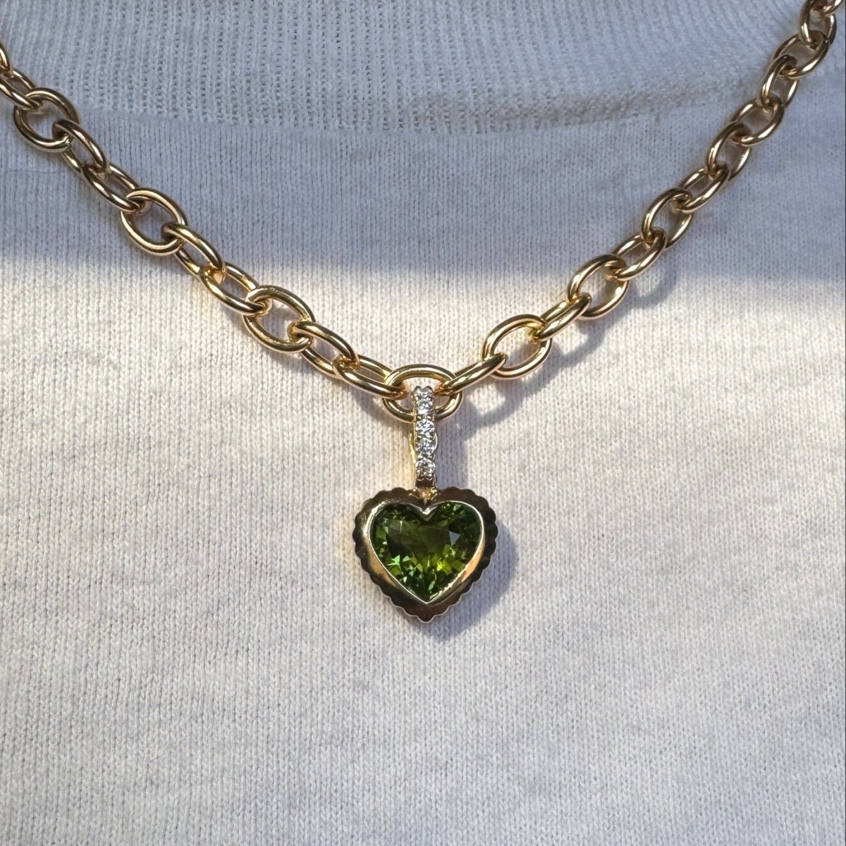 Person wearing a gold pendant with a heart-shaped green tourmaline and a diamond bail on a gold chain against a white top