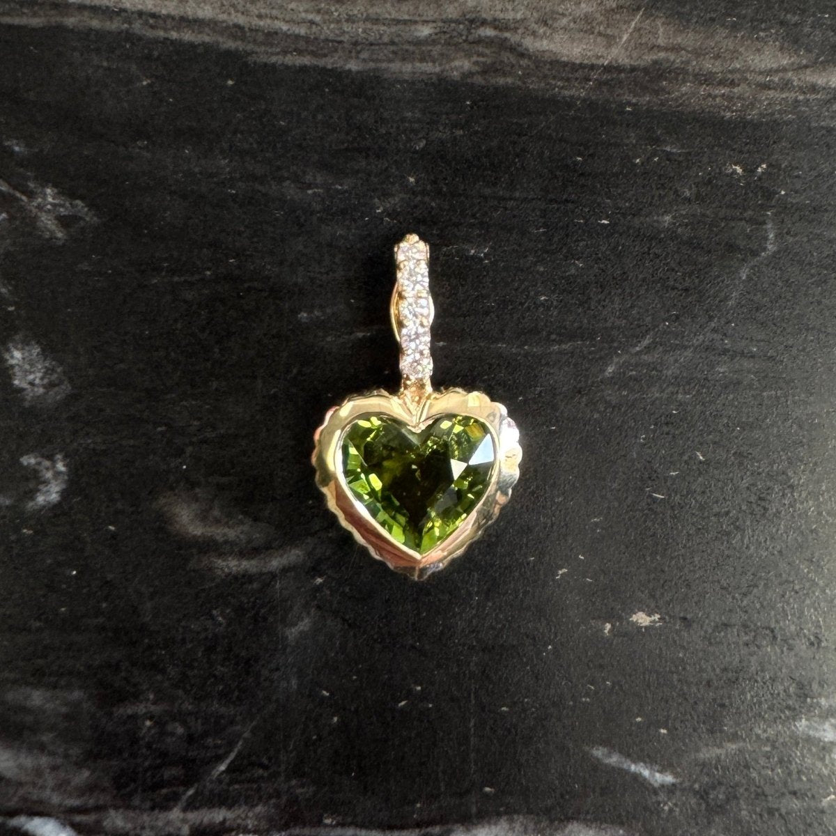 Gold pendant with a heart-shaped green tourmaline and a diamond bail against a black an white marble background 