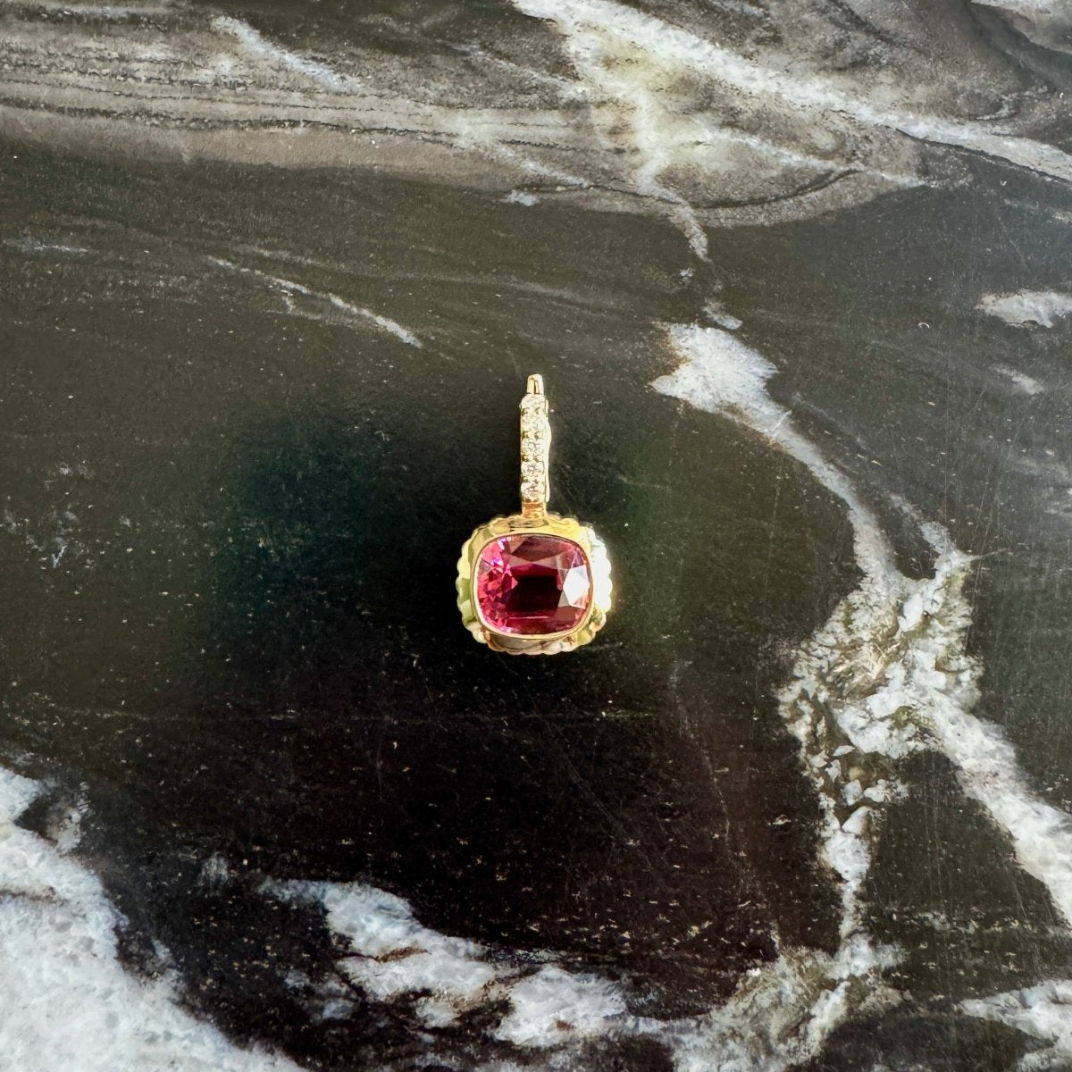 A gold pendant with a cushion-cut red gemstone, displayed on a dark, veined marble surface.