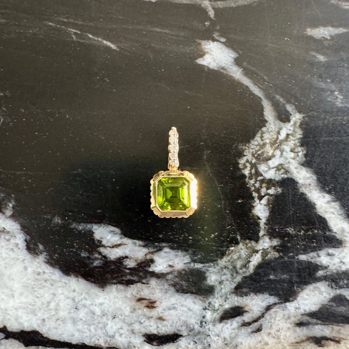 A gold pendant with a square-cut green gemstone, displayed on a black and white marbled surface.