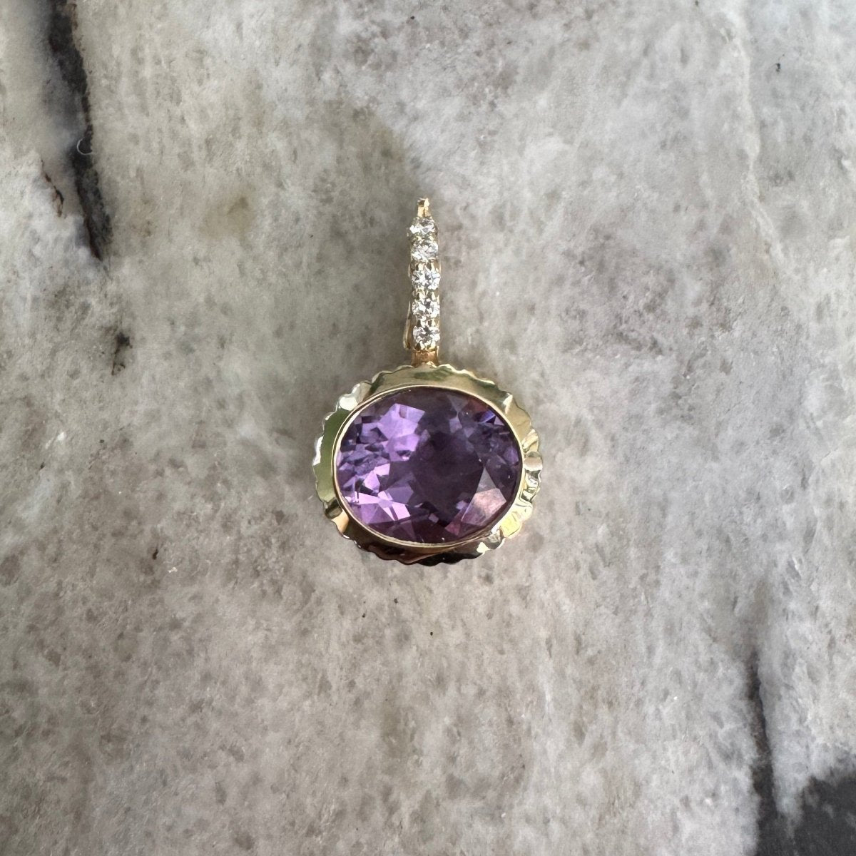 Gold pendant with an oval-cut purple amethyst and a diamond bail against a black and white marble background 