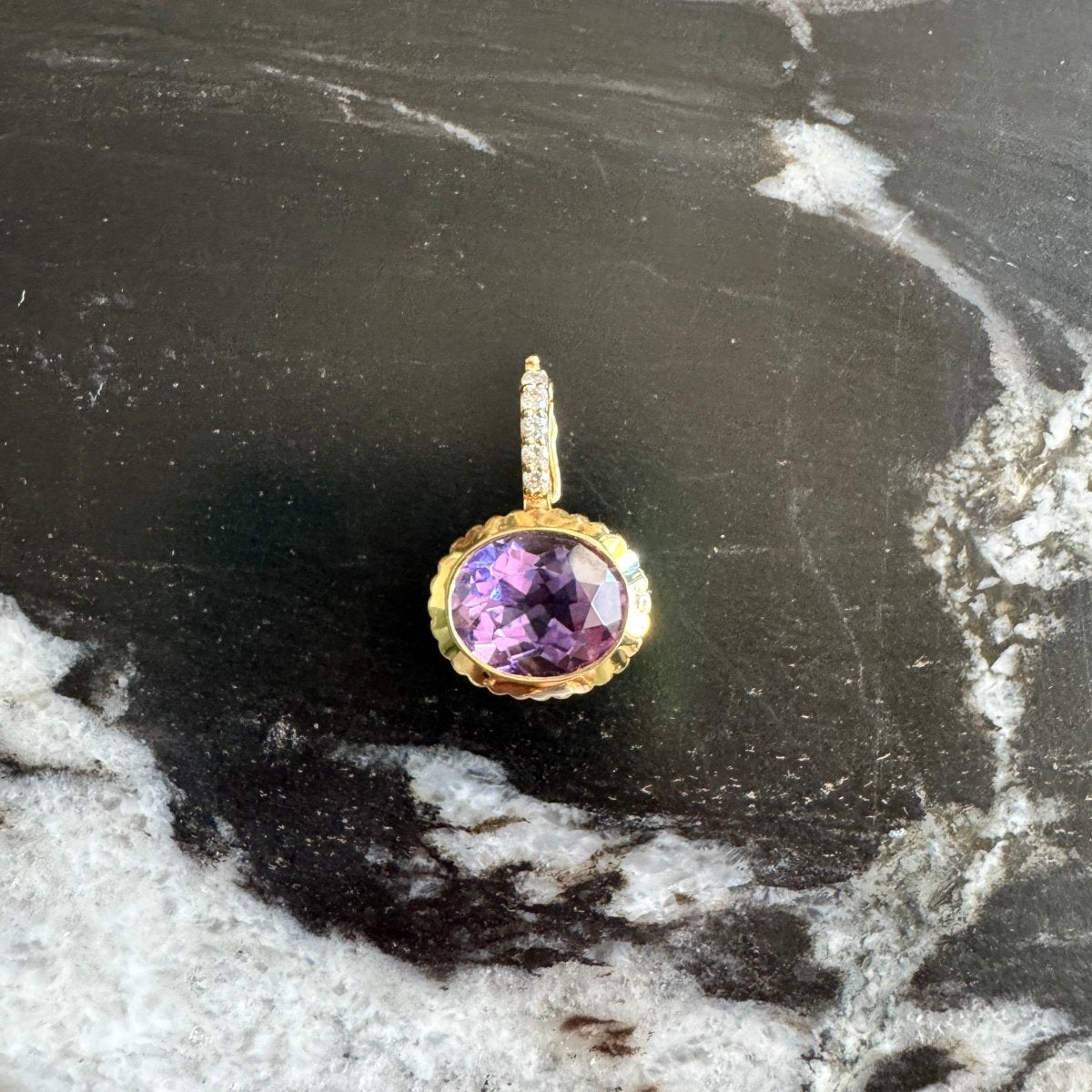 A gold charm with a round purple gemstone, displayed on a black and white marbled surface.