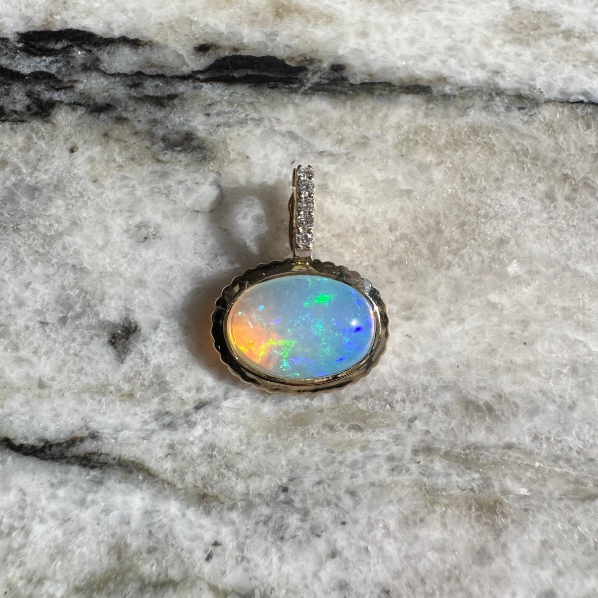 Gold pendant with an oval multicolored opal and a diamond bail against a black and white marble background 