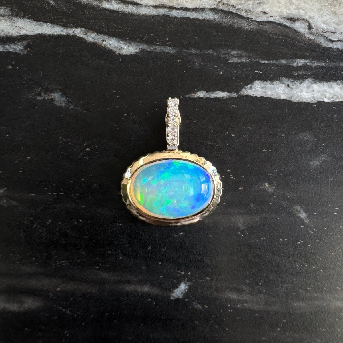 Gold pendant with an oval multicolored opal and a diamond bail against a black and white marble background 