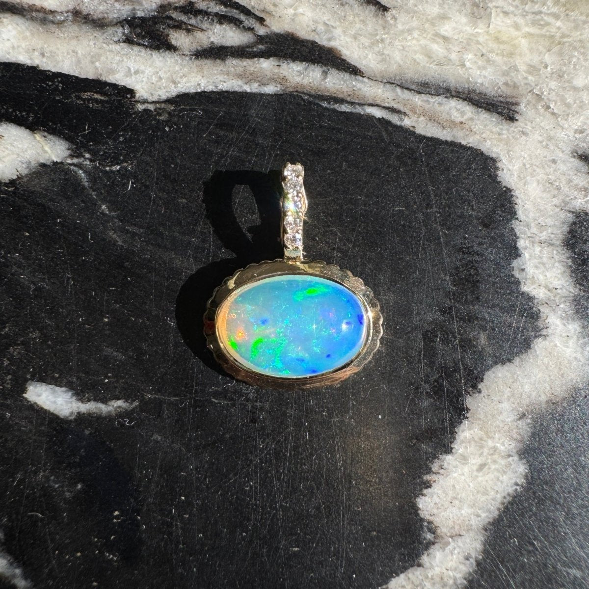 Gold pendant with an oval multicolored opal and a diamond bail against a black and white marble background 