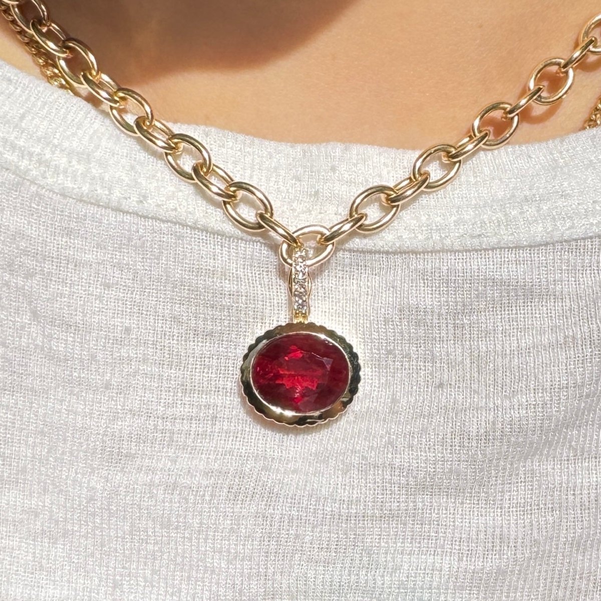 Person wearing a gold pendant with a red sunstone and a diamond bail on a gold chain against a white top