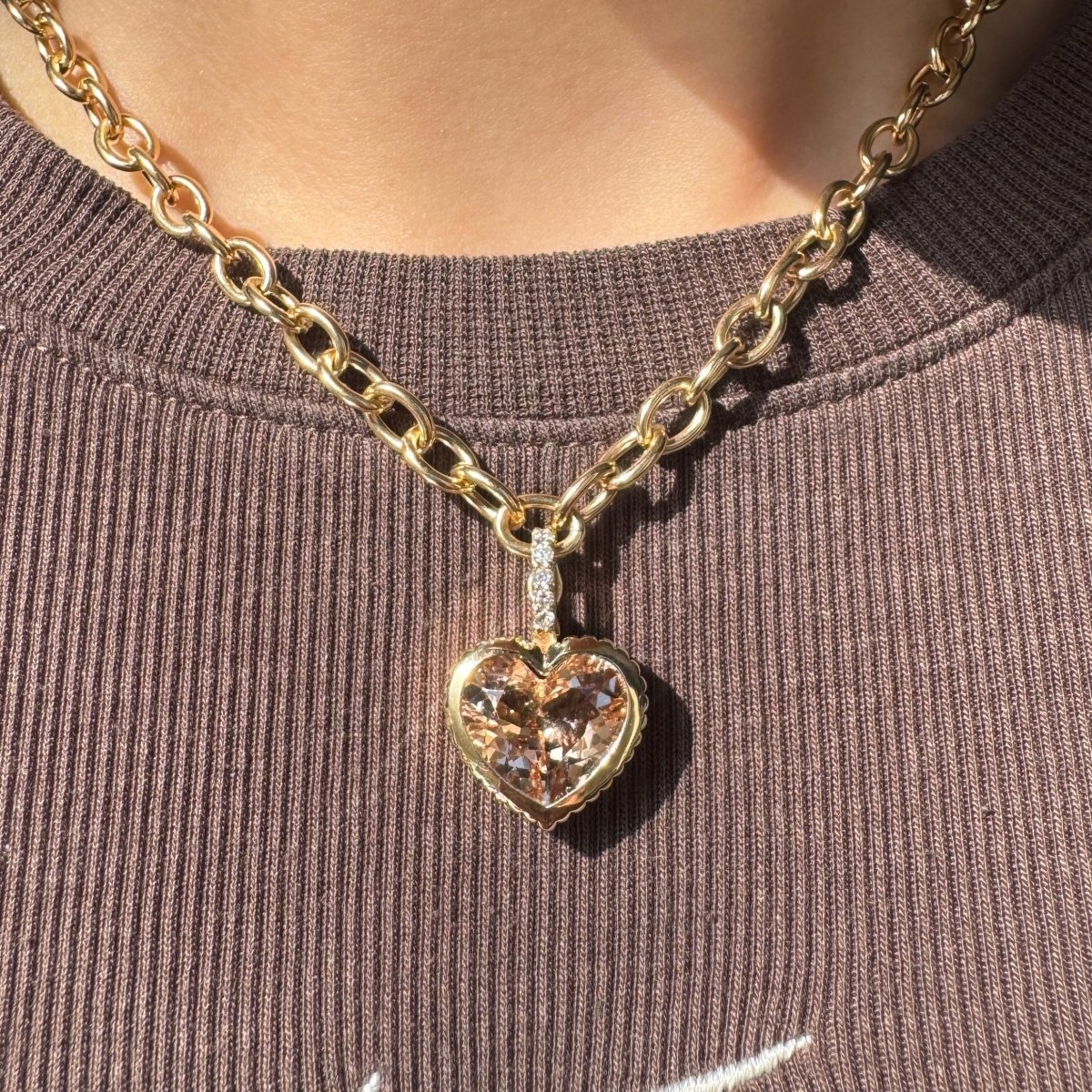 Person wearing a gold pendant with a peachy heart-shaped morganite and a diamond bail against a brown top 