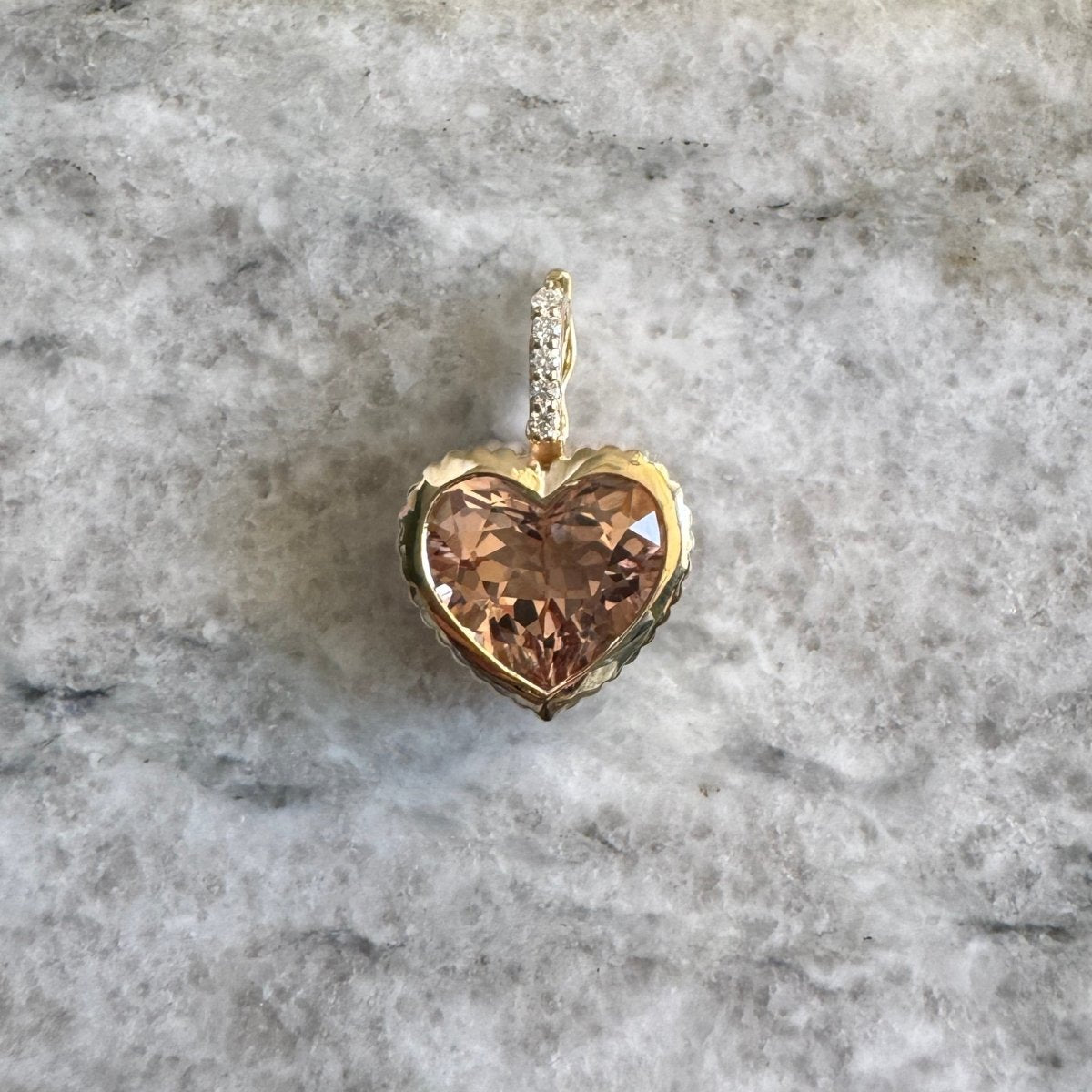 Gold pendant with a heart-shaped peachy morganite and a diamond bail against a black and white marble background