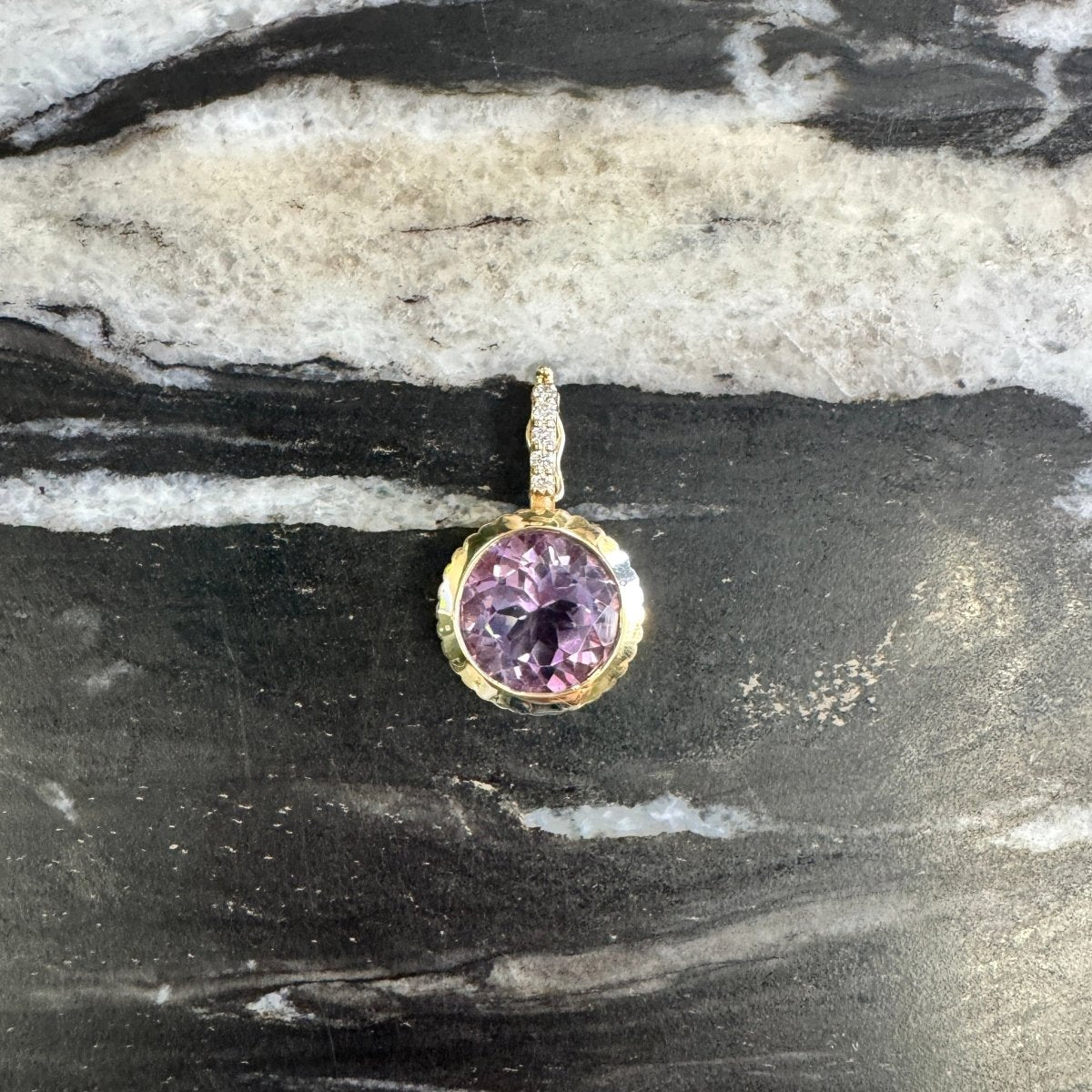 A small gold pendant with a round purple gemstone, resting on a black and white marbled surface.