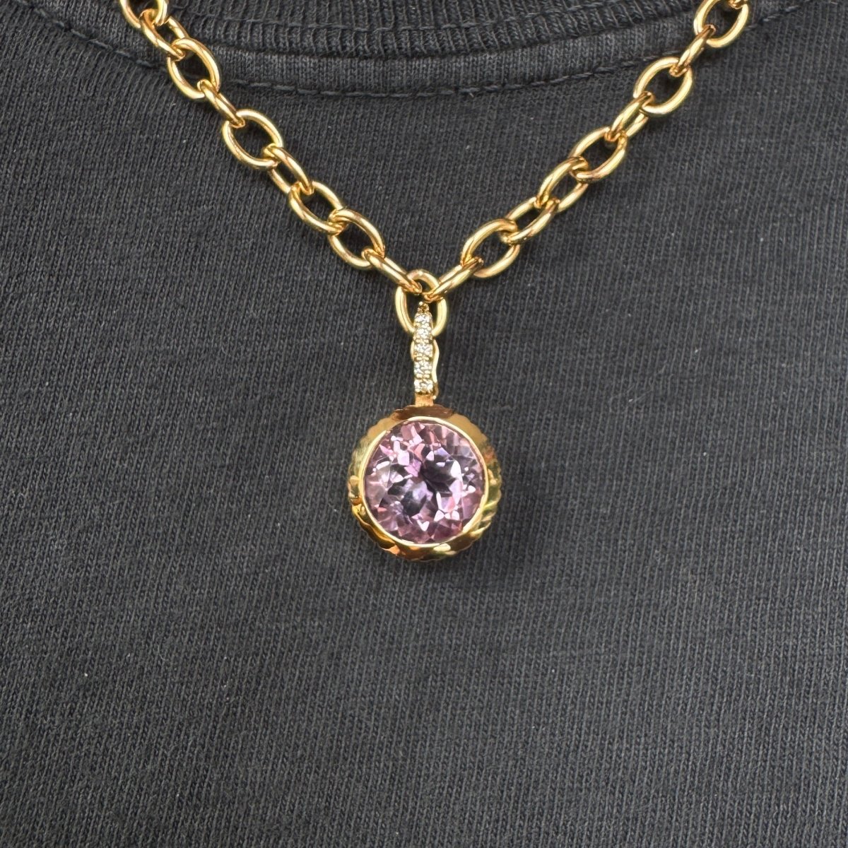 Person wearing a gold pendant with a round purple amethyst and a diamond bail on a gold chain against a black top