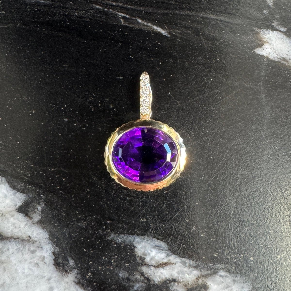 Gold pendant with an oval purple amethyst and a diamond bail against a black and white marble background
