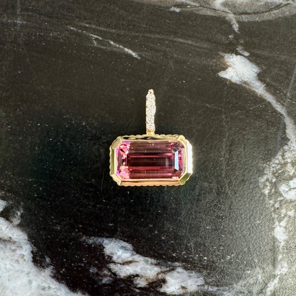 Gold pendant with a pink tourmaline and a diamond bail against a black and white marble background 