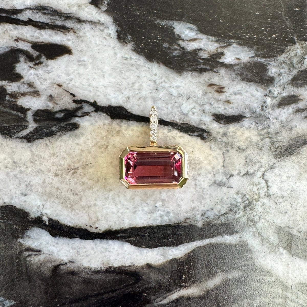 Gold pendant with a pink tourmaline and a diamond bail against a black and white marble background 