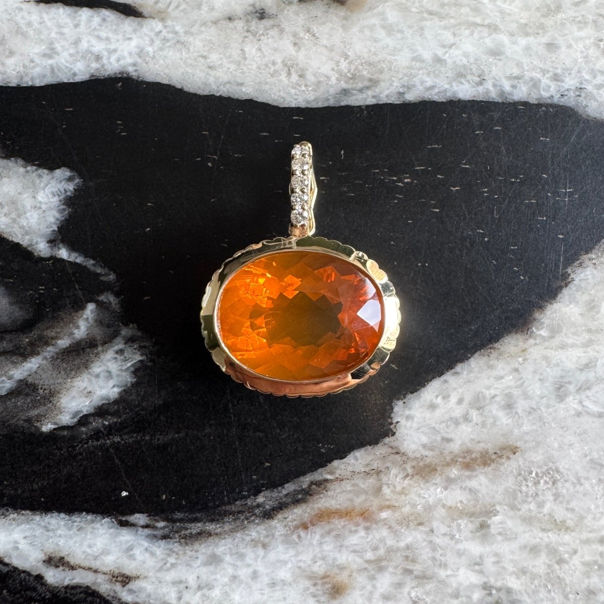 Gold pendant with an oval-cut fire opal and diamond bail against a black and white marble background