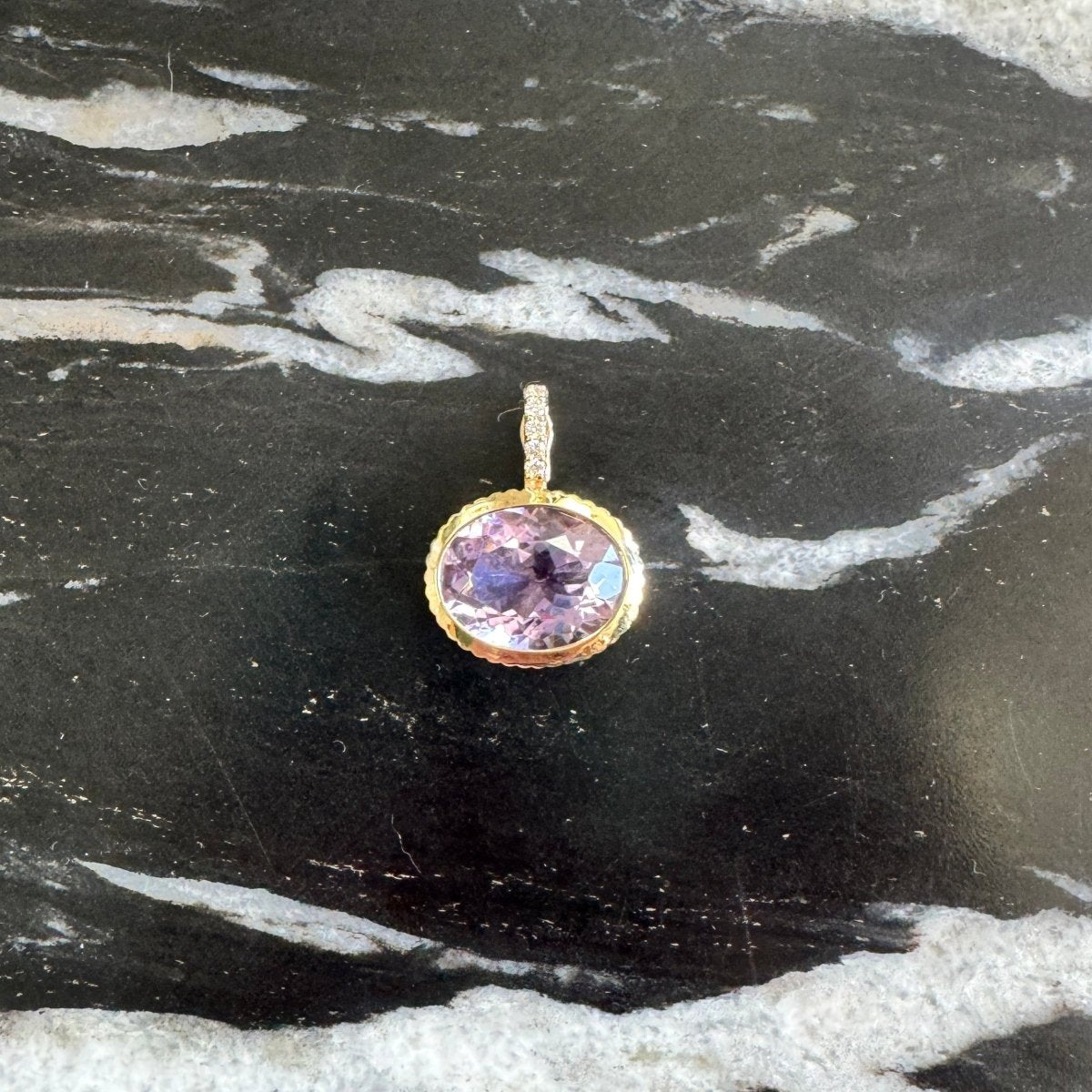 A small oval pendant with a purple gemstone in a gold bezel setting, placed on a black and white marbled surface.