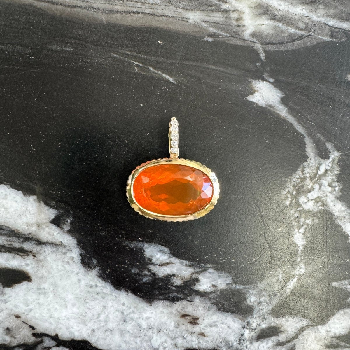 Gold pendant with an oval orange fire opal and a diamond bail on a black and white marble background