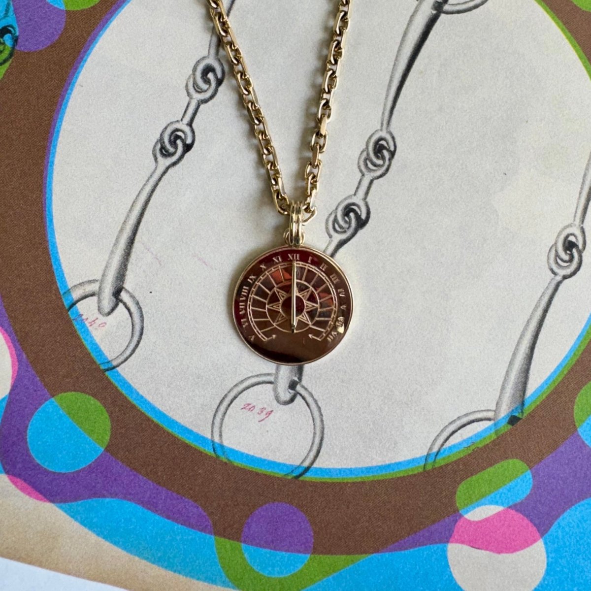 A gold chain necklace with a round pendant featuring a red and gold compass design, displayed on a colorful vintage-style illustrated card.