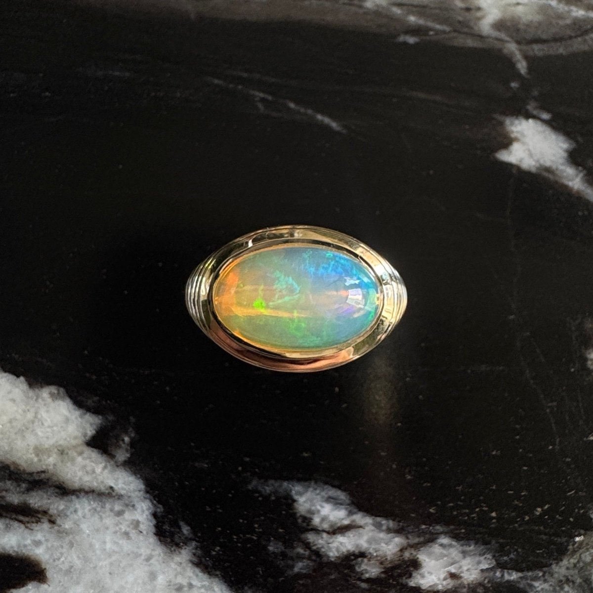 Gold ring with an oval cabochon rainbow opal against a black and white marble background