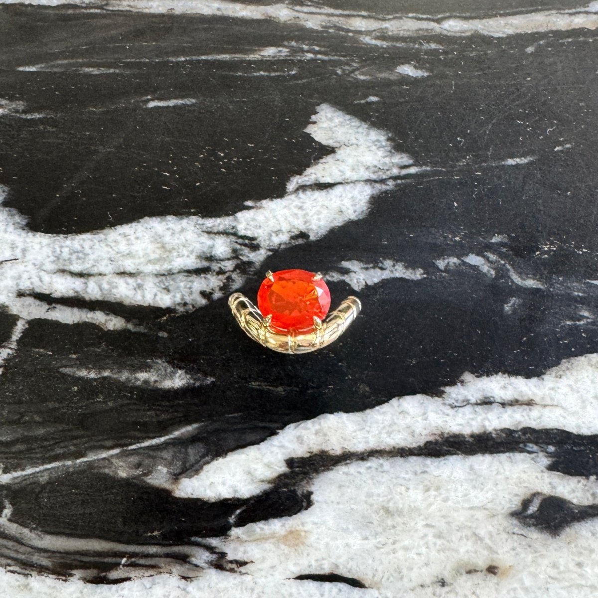 A small gold ringwith a round orange-red gemstone, displayed on a black and white marbled surface.