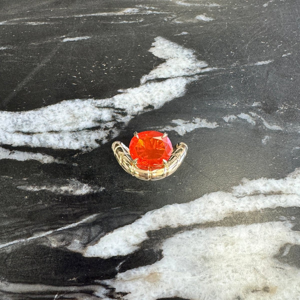 A small gold  ring with a round orange-red gemstone, displayed on a black and white marble surface.