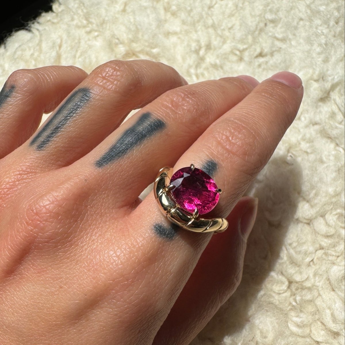 Hand wearing a gold ring with oval-cut rubellite and a curved band against a cream background
