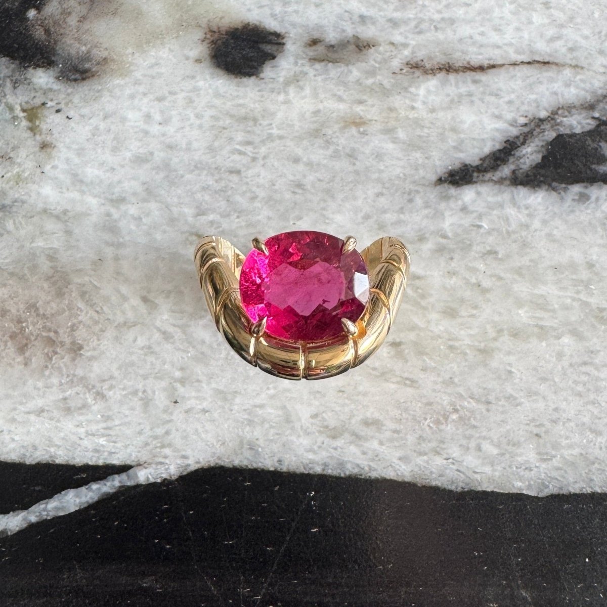 Gold ring with oval-cut rubellite and a curved band against a black and white marble background 