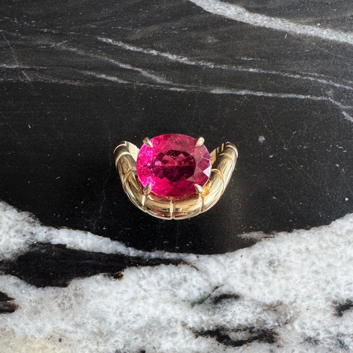 Gold ring with oval-cut rubellite and a curved band against a black and white marble background 