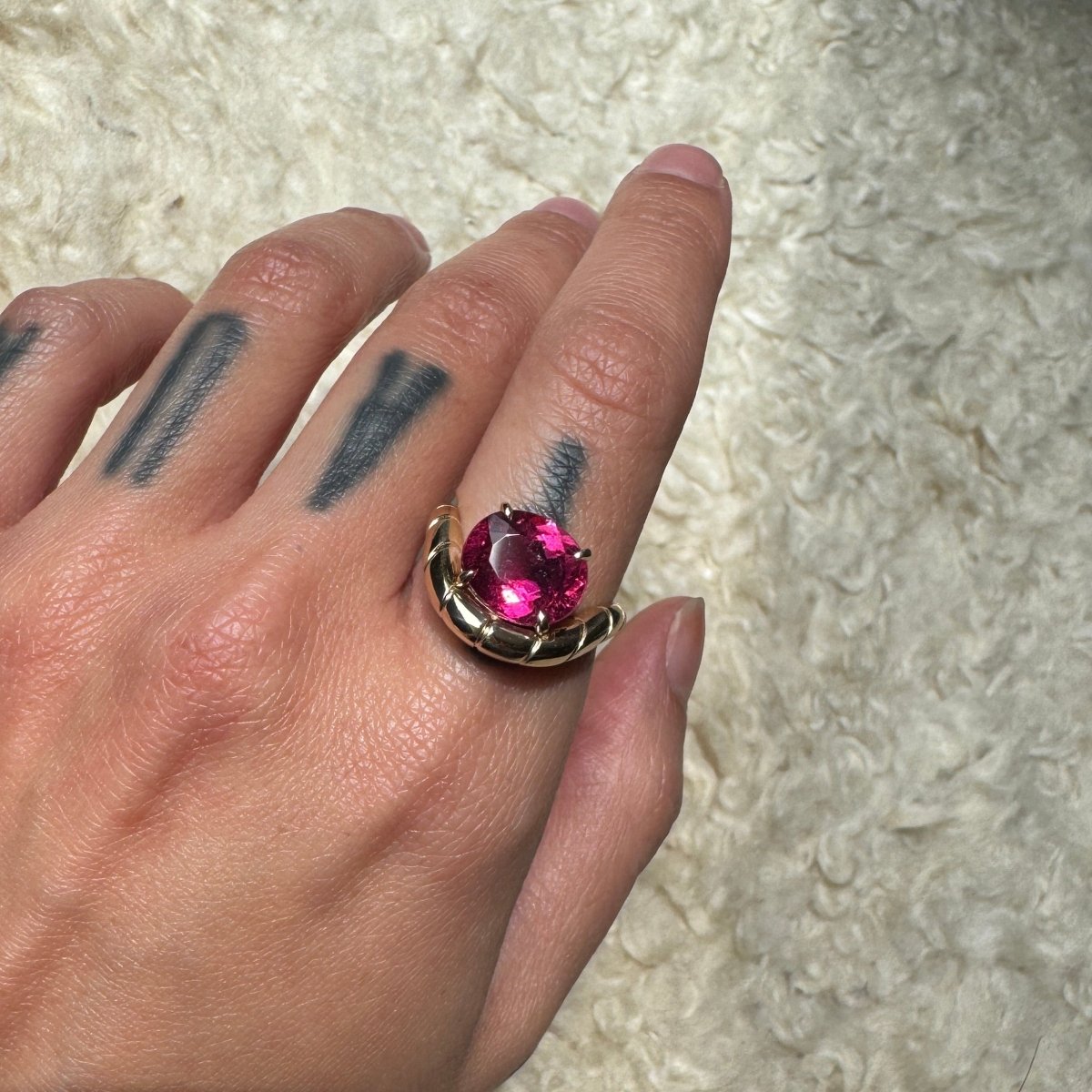 Hand wearing a gold ring with oval-cut rubellite and a curved band against a cream background