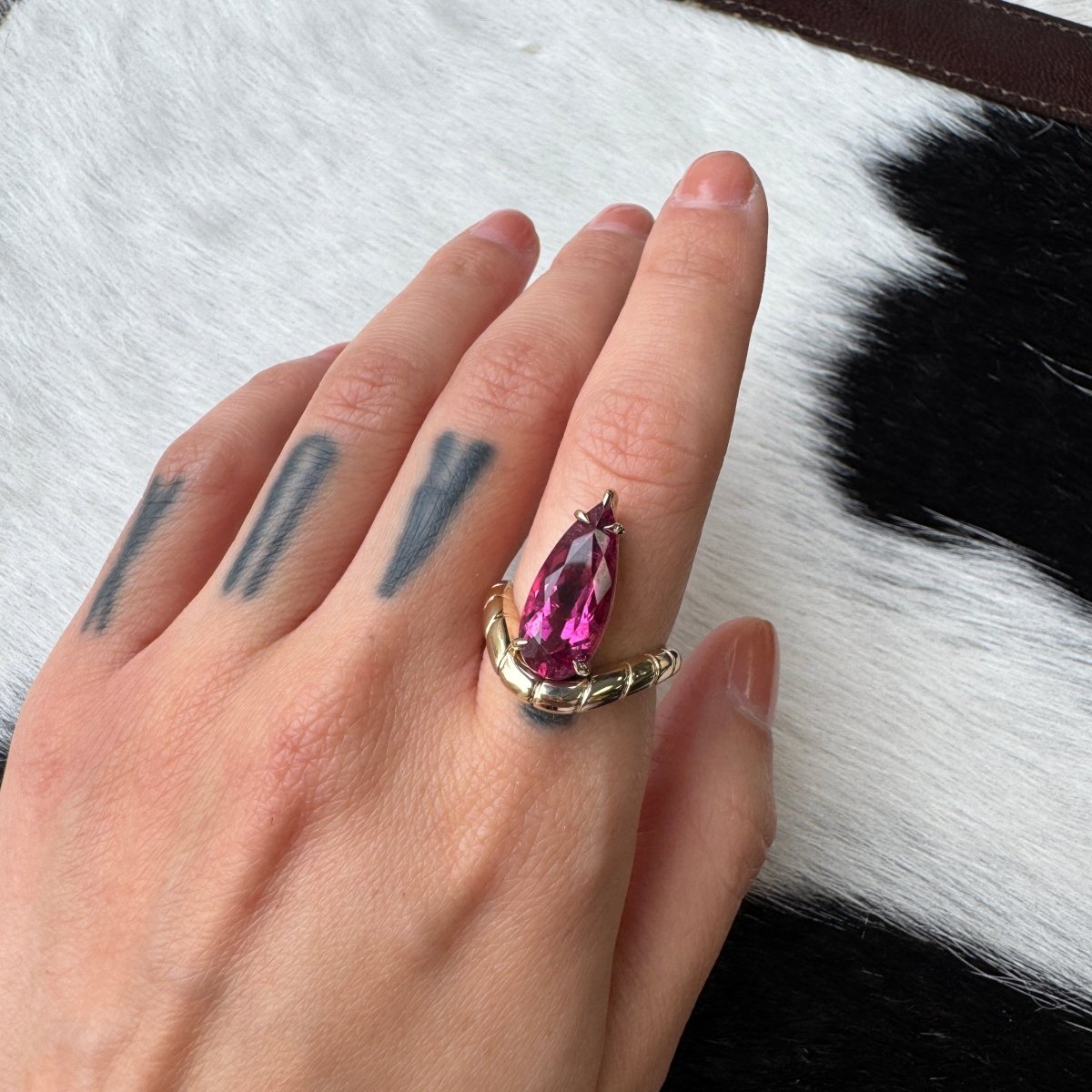 Hand wearing a gold ring with a curved band and a pear-shaped rubellite against a black and cow print background