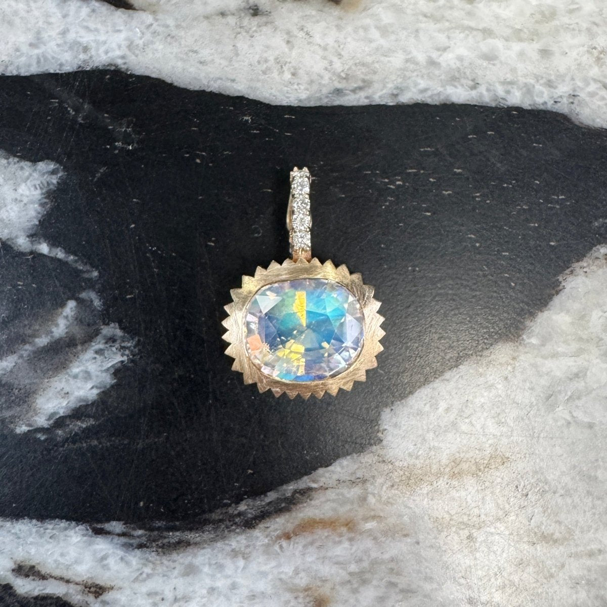Gold pendant with a multicolored cushion cut Moonstone and a diamond bail against a black and white marble background 