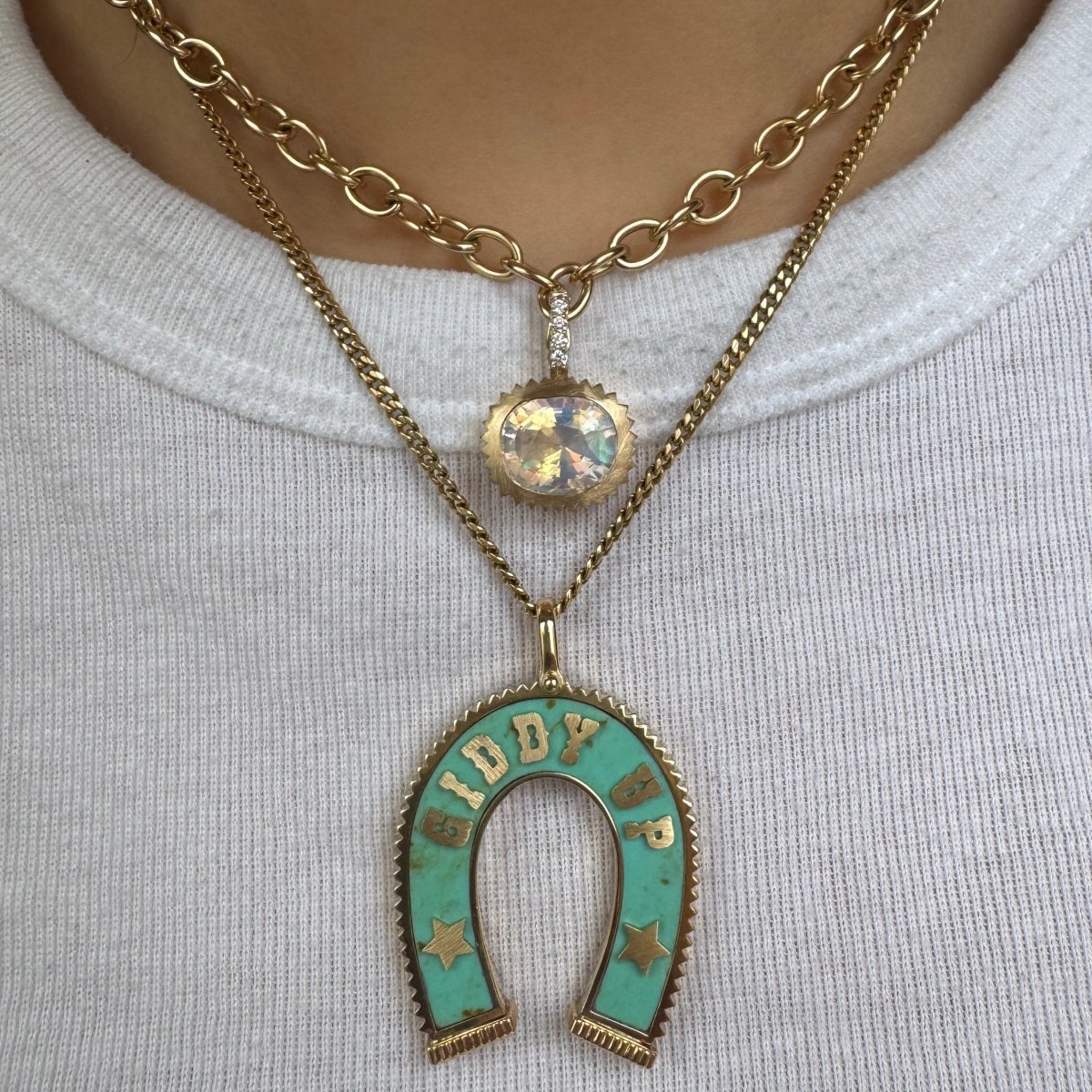 Person wearing multiple gold pendants and necklaces including a Gold pendant with a multicolored cushion cut Moonstone and a diamond bail against a a white top