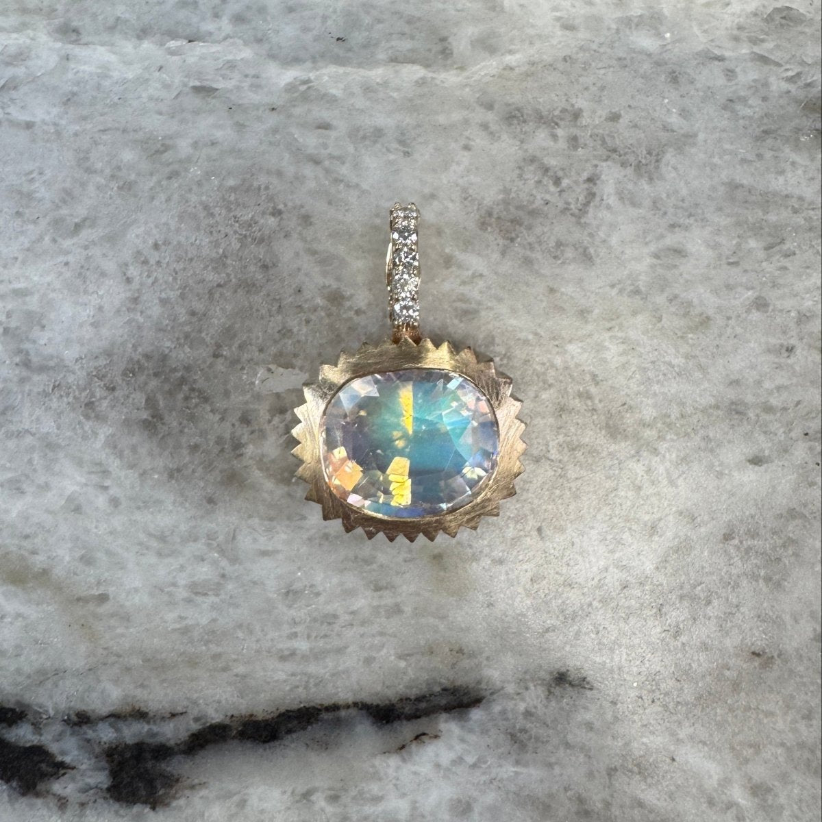 Gold pendant with a multicolored cushion cut Moonstone and a diamond bail against a black and white marble 
